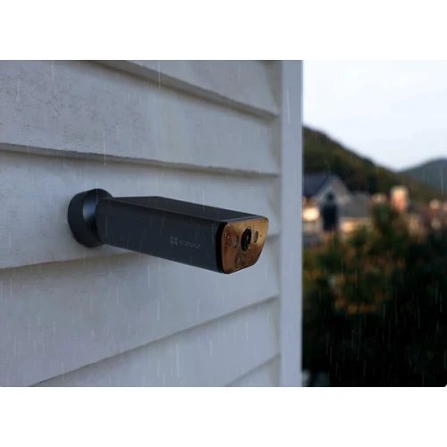 Kamera monitoringu zamontowana na zewnętrznej ścianie budynku podczas deszczu. W tle widać rozmyty krajobraz z drzewami i budynkami.